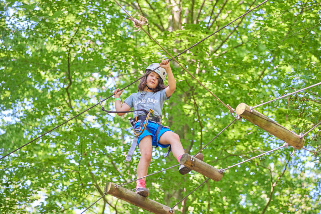 Jongetje met witte helm klimt over brug met houten palen als treden en kijkt in de camera.