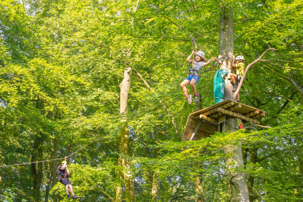 Jongetje springt van het platform met een hand in de lucht en gaat van de zipline. op de achtergrond is een man ook van de zipline aan het gaan.