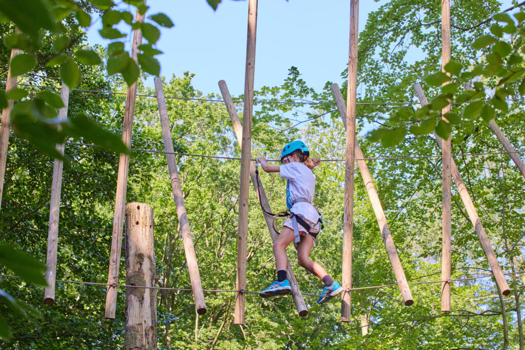 Meisje met blauwe helm klimt over touw tussen diagonale houten palen.