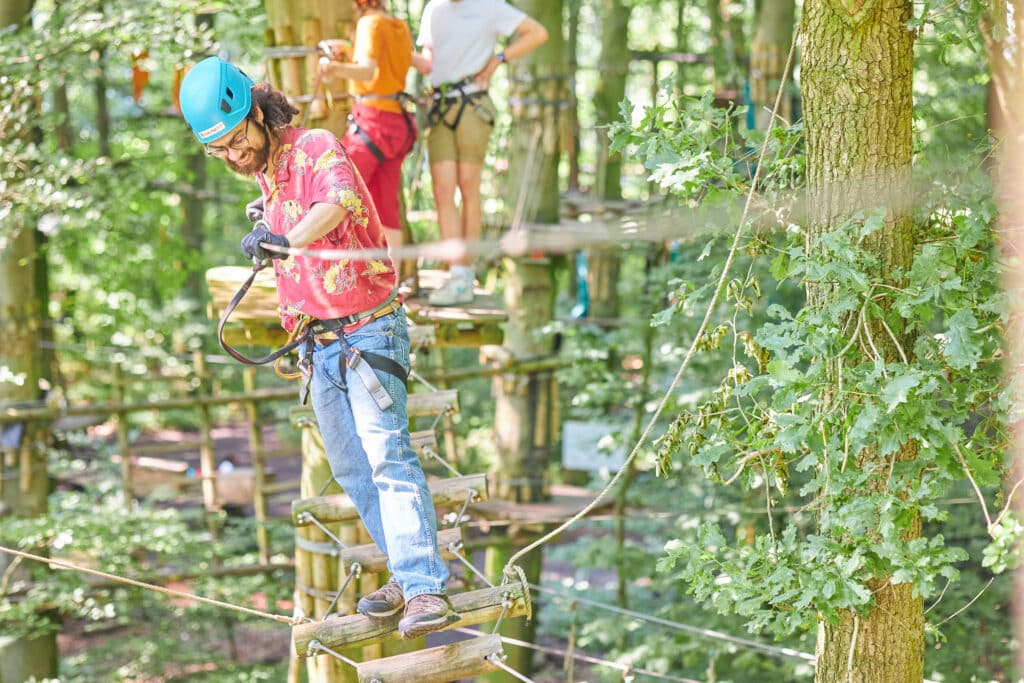 Man in red blouse and blue helmet climbs a bridge with wooden posts.