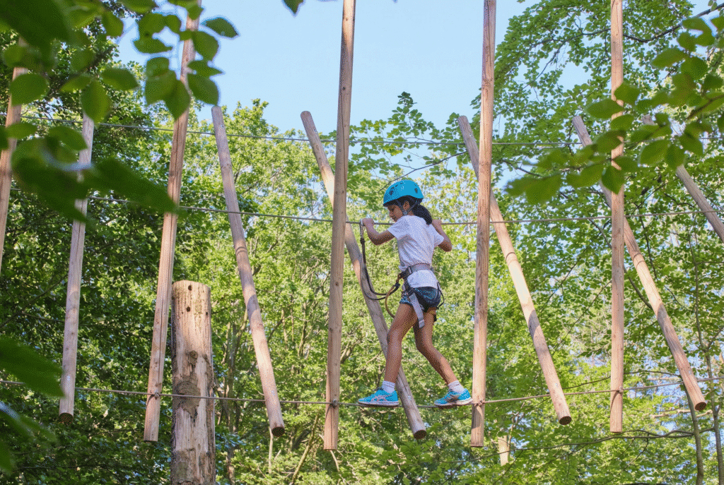 Mädchen mit blauem Helm klettert über ein Seil zwischen diagonalen Holzpfosten.