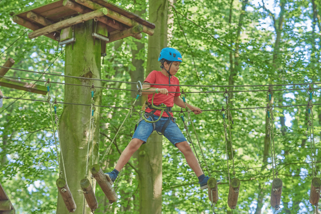 Jongen met blauwe helm en rood shirt staat op twee balken van een brug. de balken onder hem bewegen uit elkaar.