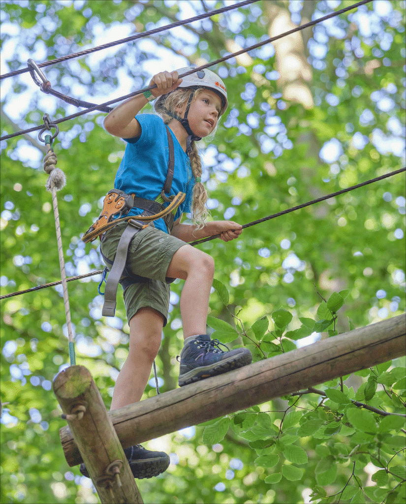 Meisje met een vlecht en een witte helm klimt over een houten balk. Ze kijkt geconcentreerd naar voren.